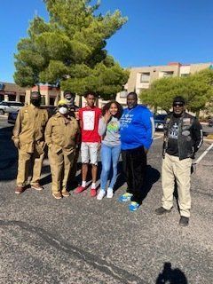 Group of people posing outdoors in parking lot. Sunny day. Several wearing masks, casual clothing.