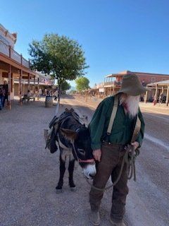 Man in Western attire leads a donkey down a dusty street in an Old West town.