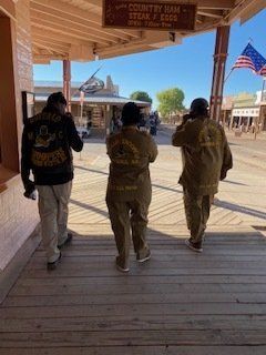 Three people in matching jackets walk on a wooden boardwalk in a western-themed town, flags in the background.