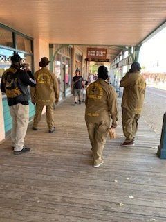 Group of people in matching tan outfits walking on a wooden boardwalk in front of shops.