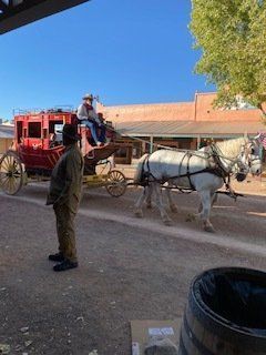Stagecoach with white horses in front of a western-style building; a person stands nearby.