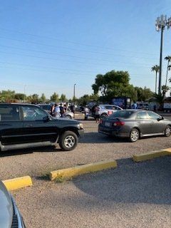 Cars parked in a gravel lot, people gathered nearby. Outdoor setting, blue sky.