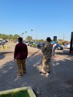 Two people stand watching an event outdoors. Cars, motorcycles, and trees are visible under a blue sky.
