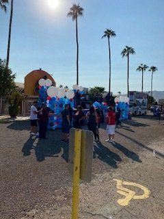 People gathering outdoors near balloons, palm trees, and a building. Sunshine.