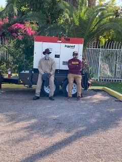 Two men in front of a generator on a trailer. One wears a mask and work clothes. The other, a t-shirt and khakis.