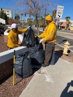 Two people in yellow hoodies collecting trash bags on a sidewalk.
