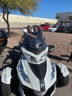 Two people smiling on a white and black three-wheeled motorcycle in a parking lot.