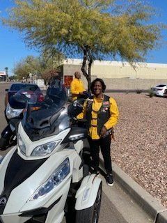 Woman on white trike motorcycle, wearing yellow shirt and black vest, parked on street. Another person in background.