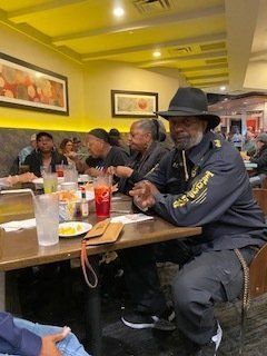 People seated at a restaurant table, one man in a hat and jacket, eating and conversing.