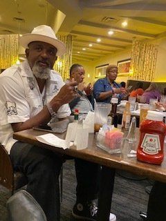 Man in white shirt and hat sits at a restaurant table, looking towards the camera. Other people dine nearby.