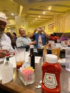 People seated at a restaurant table. One person takes a photo. Ketchup bottle in foreground.