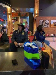 Two people seated at a bowling alley. One in a dark uniform, cowboy hat; one in a blue jacket. Party supplies on the table.