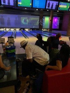 People bowling in a dimly lit alley with screens displaying images. One person wearing a white shirt and hat is seated.
