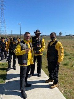 Three men in yellow and black vests pose on a sunny day. A group of people are behind them.
