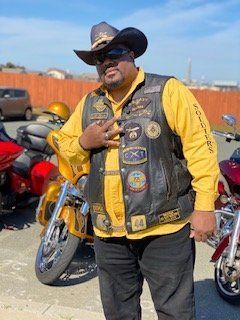 Man in cowboy hat and biker vest, standing near motorcycles. He is making a hand gesture.