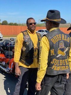 Two men in yellow shirts and leather vests with motorcycle, one with hat, outdoors.