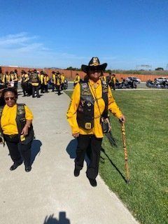 People in yellow and black vests and jackets walking outdoors. One person uses a cane. Motorcycles in the background.