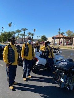 Three motorcyclists in yellow jackets and vests stand near their bikes in a sunny parking lot.