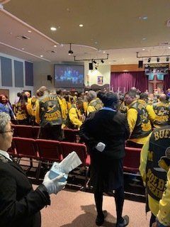 People in yellow vests with patches stand inside a large auditorium. A woman in a black outfit holds papers and looks toward the crowd.