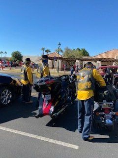 Motorcycle riders in yellow vests stand near their bikes in a parking lot. Buildings and cars are in the background.