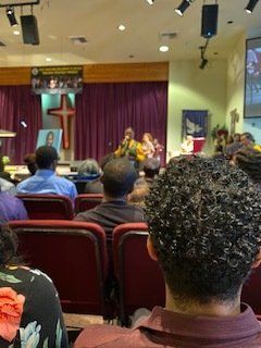 People seated in a church listening to speakers on a stage with a cross backdrop.