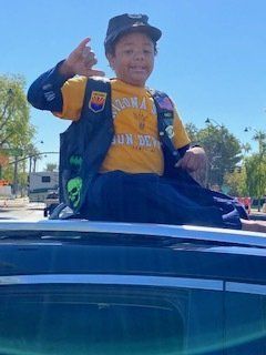 Child in yellow shirt and vest, sitting on car roof, giving a shaka sign.