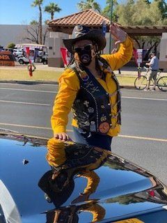 Woman in cowboy hat and vest waves, reflected on car hood, outdoors.