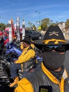 Two people in military-style hats and jackets on a motorcycle, outdoor setting, possibly a parade.