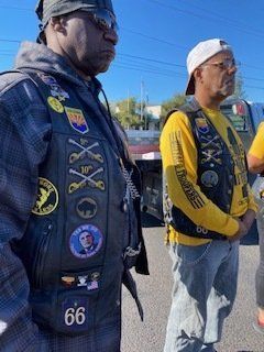 Two men in vests with patches standing outdoors. One wears a bandana, the other a baseball cap. Both have serious expressions.