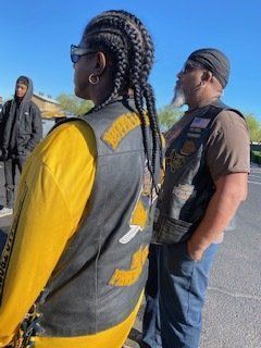 Two people wearing biker vests stand outdoors. One has braids, the other a goatee. A person in a black hoodie stands behind them.