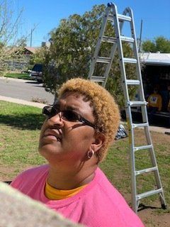 Woman in pink shirt and sunglasses looking up near a ladder outdoors.