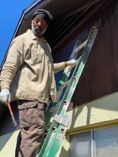 Man on ladder painting brown wood siding, wearing gloves and a cap. Yellow trim and blue sky.
