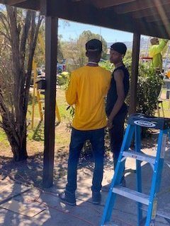 Two people in front of a structure, one in yellow shirt, the other in black. A ladder is present.