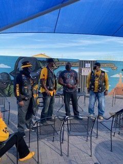 Four men in vests and hats stand outdoors near a mural and chairs, under a blue canopy.