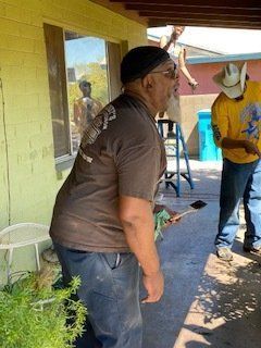 Man painting exterior; others on ladder and at work. Outdoor setting, green, yellow, and blue.