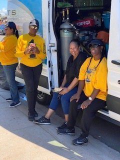 Four people in yellow shirts outside a white van. Two sit, two stand. One holds a phone.