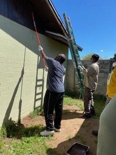 Two people painting a house trim with a roller from a ladder on a sunny day.
