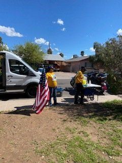 Two people near a van and table with American flag. Sunny day.