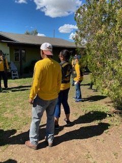 People in yellow shirts and blue jeans stand outside a house on a sunny day.
