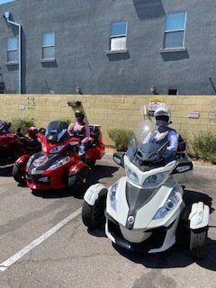 Three Can-Am Spyder motorcycles parked on asphalt; two red and one white, people riding them.