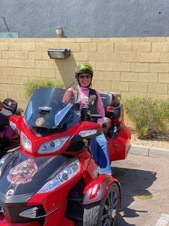 Woman on a red Can-Am Spyder motorcycle, wearing pink and blue, with a helmet, parked near a building.