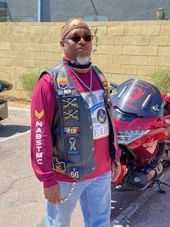 Man in biker vest stands near red motorcycle; outdoors, sunny.