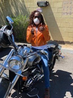 Person on a motorcycle giving thumbs up, wearing a mask and orange jacket.
