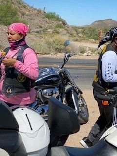 Two people in biker vests stand near a parked motorcycle on a road, desert landscape in background.