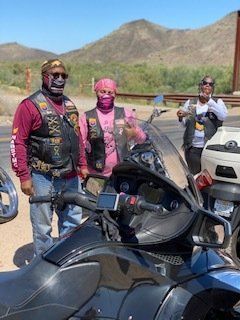 Three people in biker vests stand next to motorcycles outdoors. Mountains in the background.