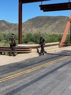 Two figures standing on a bridge, mountains in the background under a blue sky.