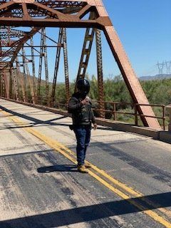 Person wearing a helmet stands on a weathered bridge with brown metal supports under a blue sky.