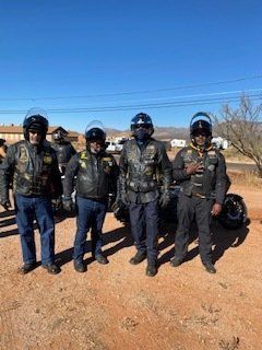 Four people in biker gear pose outdoors, helmets on, next to a motorcycle.