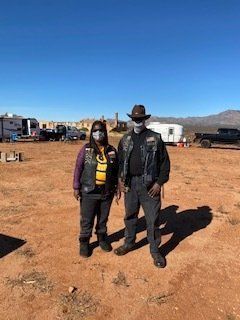 Two people in biker vests and masks stand outdoors in a desert landscape.