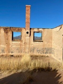 Ruined adobe building with chimney against a blue sky, mountain visible through empty window frames, and dry grass.